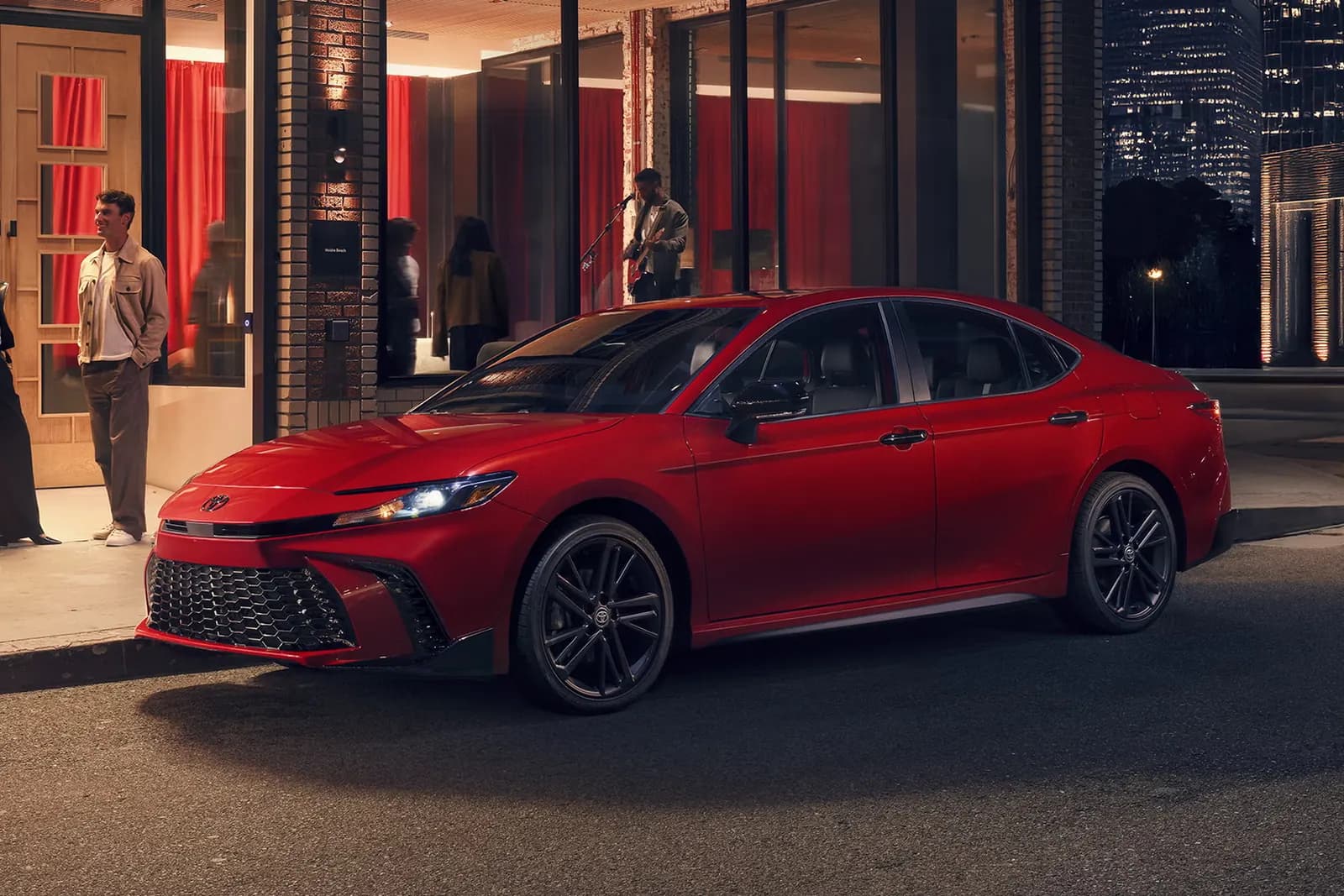 2027 Toyota Camry hybrid sedan in red, front three-quarter view, parked at night on a city street